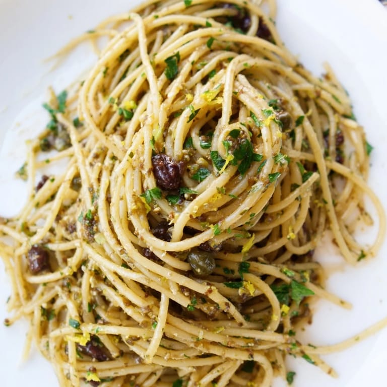 A rustic serving of Olive Tapenade Pasta in a white bowl, garnished with fresh parsley and lemon zest beside a glass of white wine.  